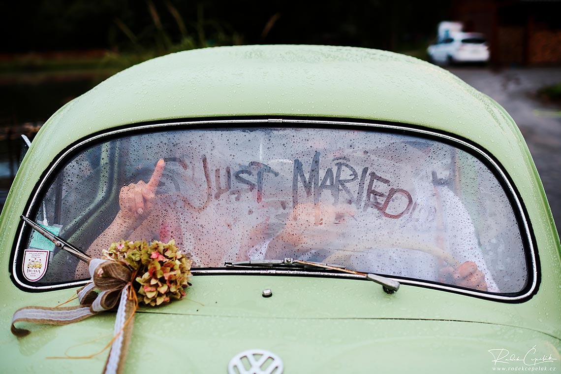 svatební fotografie novomanželů v autě za sklem s nápisem Just Married