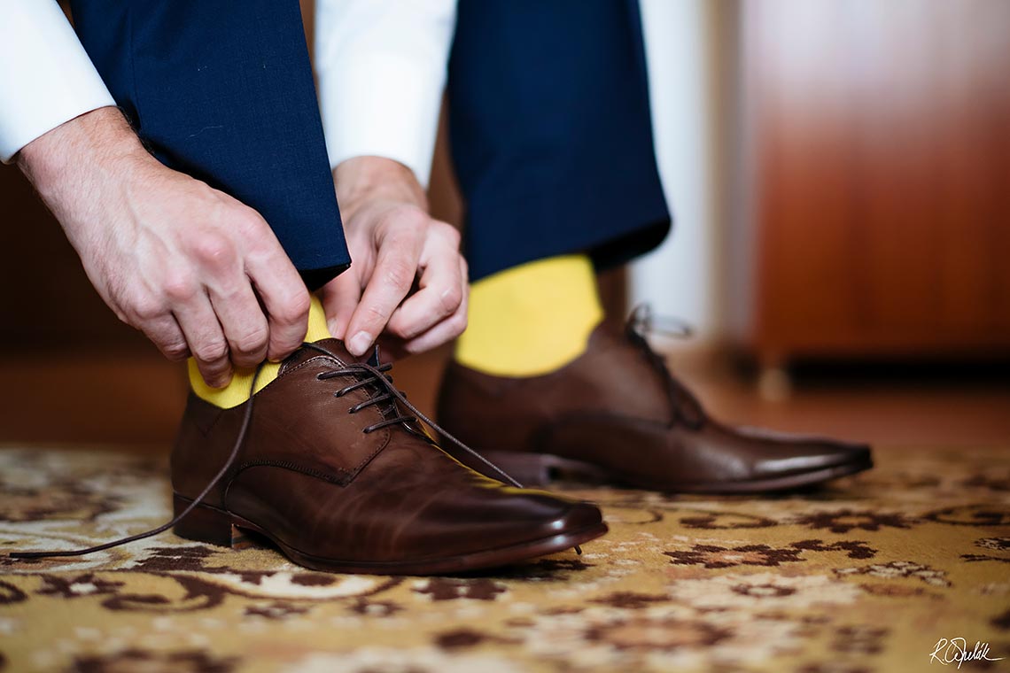 ženich si zavazuje hnědé svatební boty v kombinaci se žlutými ponožkami