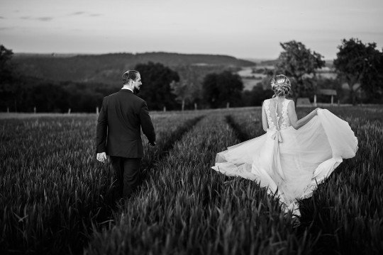 černobílá svatební fotografie nevěsty a ženicha jak prochází v poli kolejemi od traktoru